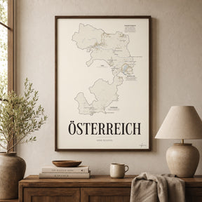 Österreich Wine Map poster by Corkframes in a dark frame above a wooden sideboard with books, a ceramic lamp and an olive branch